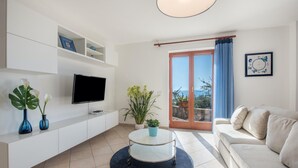 Ein modernes Wohnzimmer mit Flachbildfernseher, weißen Regalen, einem runden Couchtisch, einer Couch und Blick ins Freie durch eine Glastür.