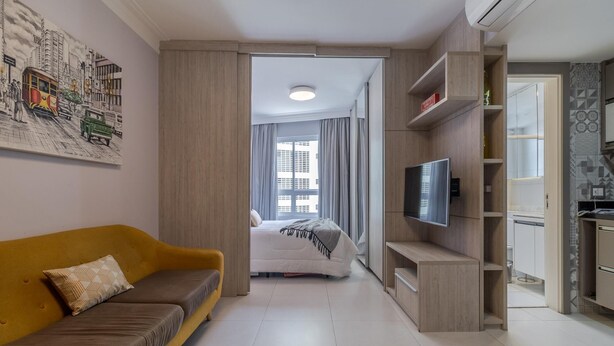 A modern living room with a yellow sofa, a flat-screen TV mounted on a wooden wall, and a kitchen area with a countertop and cabinets. - Av0703 Studio Avanhandava (São Paulo)
