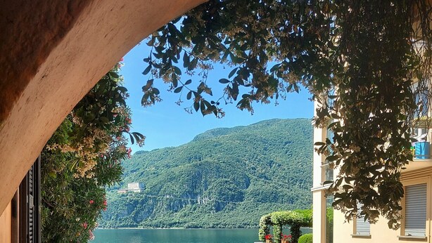 A view through an archway overlooking a lake with boats docked, a gravel path, and a building with a stone wall. - Our Happy Place Detached vacation home with garden (Mandello del Lario)