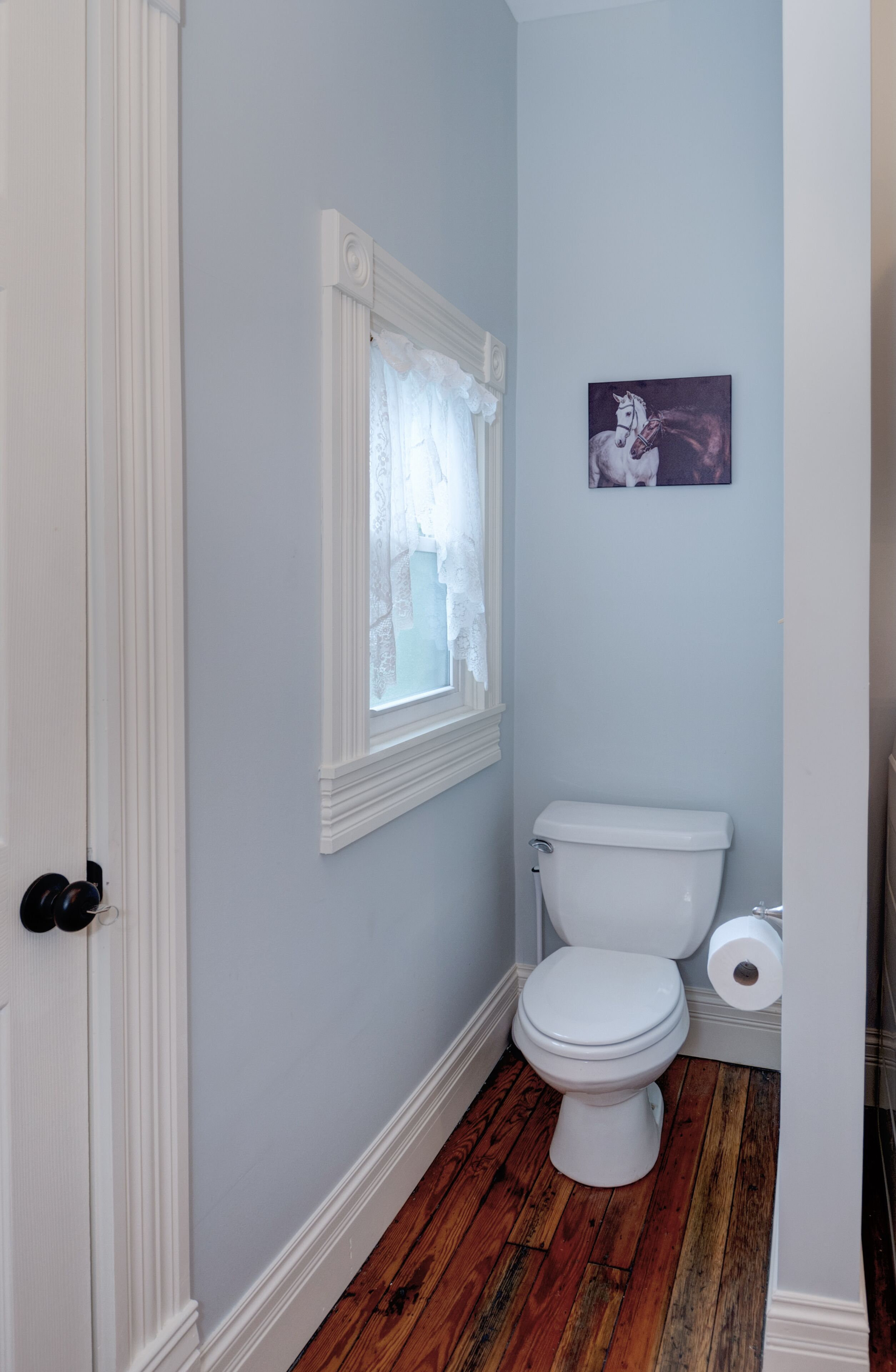 Un baño con inodoro blanco, una ventana con cortinas de encaje y un cuadro de un caballo en la pared.
