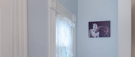 Un baño con inodoro blanco, una ventana con cortinas de encaje y un cuadro de un caballo en la pared.