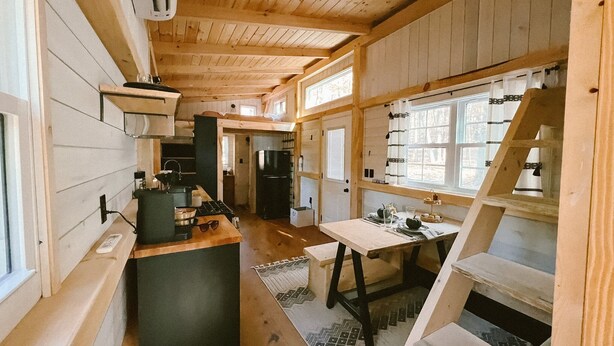 A compact kitchen with a stove, sink, and refrigerator. There is a dining area with a table and chairs. The room has wooden walls and a sloped ceiling. - COZY PSU Rustic Tiny Home and Fishing Retreat (Petersburg)