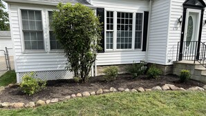 Une maison blanche avec un jardin bien entretenu, comprenant un petit arbre et une bordure de pierres.