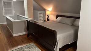 Une chambre avec un grand lit, une table de chevet avec une lampe, une étagère blanche et une petite fenêtre avec des stores.