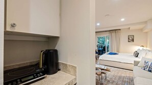 Eine Küchenzeile mit Kaffeemaschine und digitaler Waage, ein Bett mit blauem Handtuch und Blick auf einen Balkon mit Gartenmöbeln.
