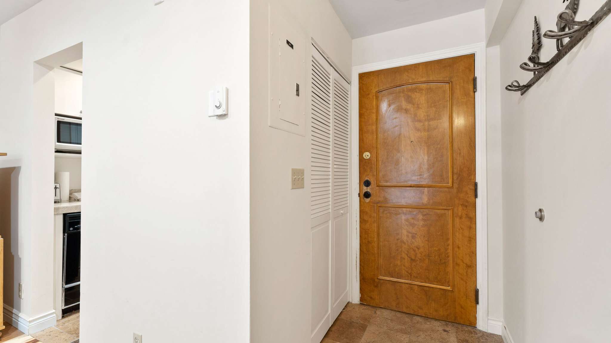 Un couloir avec une porte en bois, un mur blanc, un thermostat et une grille d’aération.