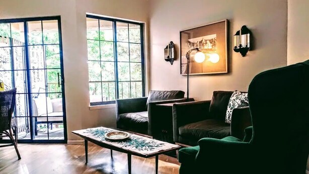 Een woonkamer met een houten vloer, een groene fauteuil, een zwarte bank en een tafel met een decoratief dienblad. Twee ramen bieden uitzicht op groen.
