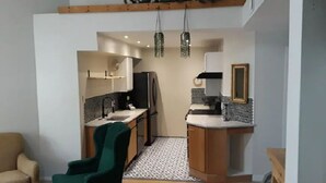 Een praktische keuken met een spoelbak, koelkast en een klein aanrecht. Een stoel staat op de voorgrond en er hangt een spiegel aan de muur.