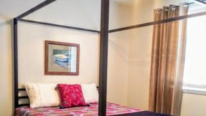 Een hemelbed met een kleurrijk beddegoed, een ingelijst kunstwerk aan de muur en een raam met gordijnen.