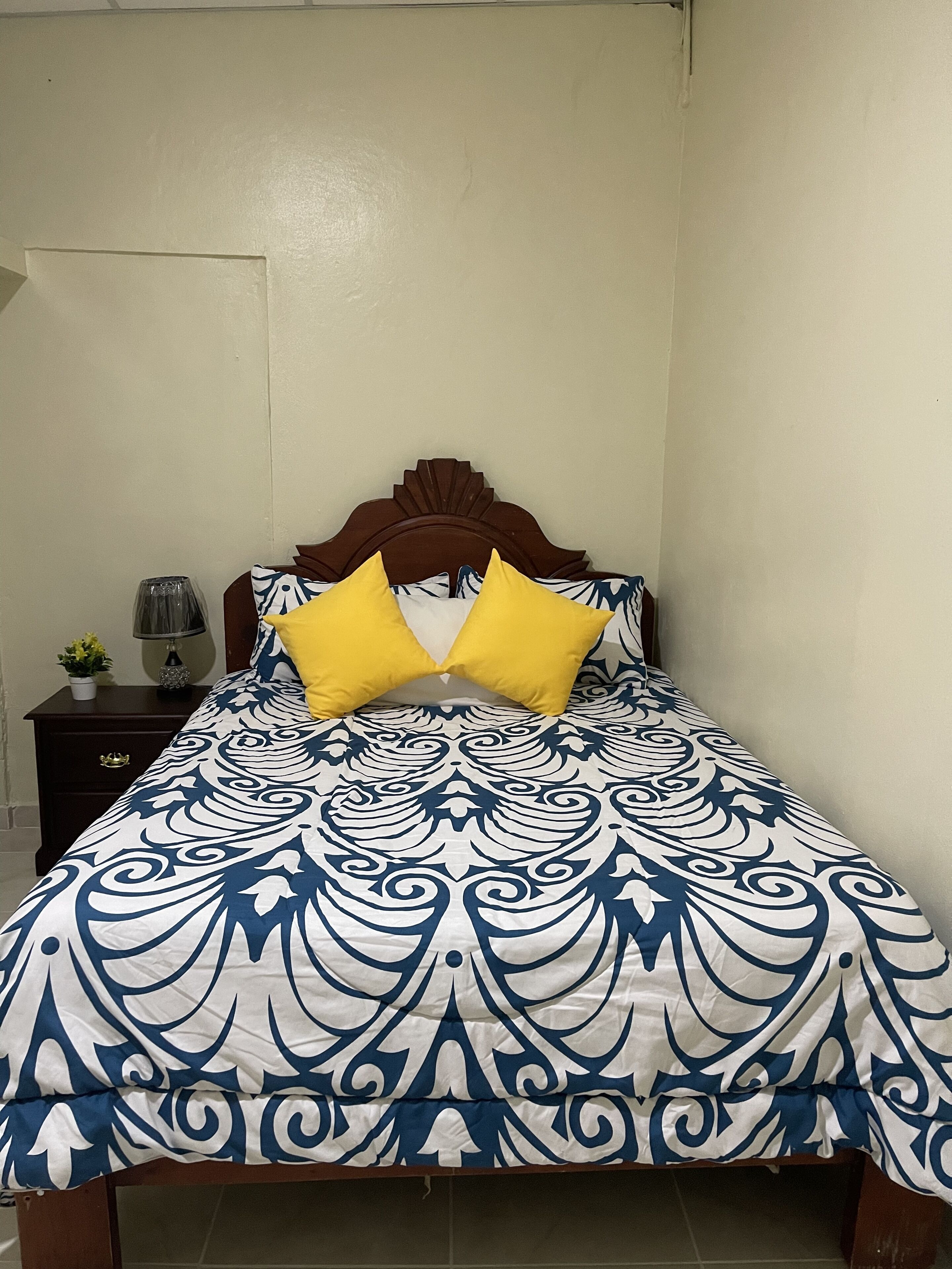 Cama com colcha estampada e duas almofadas amarelas, cabeceira de madeira e mesa de cabeceira com luminária e planta.
