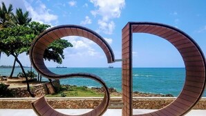 Two large, curved metal structures resembling the letter 'D' in a coastal setting with a view of the sea and palm trees. - VBermor Hotel (Santo Domingo)