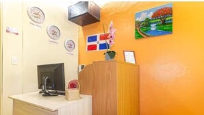 A reception area with a wooden desk, a computer, a flag, and a painting on the wall. - VBermor Hotel (Santo Domingo)