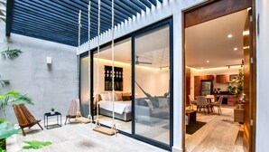 Un interior moderno con un amplio ventanal corredera que da a un dormitorio con cama, sofá y zona de comedor con mesa y sillas. Hay dos columpios afuera de la puerta.