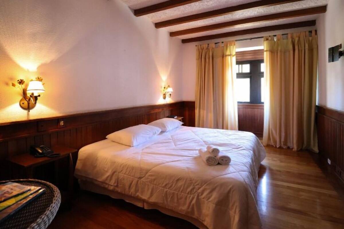Une chambre d’hôtel avec un grand lit, une tête de lit en bois, deux lampes fixées au mur et une fenêtre avec des rideaux.