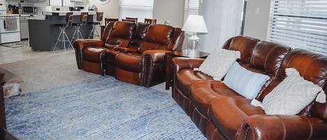 A living room with a brown leather sofa set, a blue patterned rug, and a kitchen in the background.