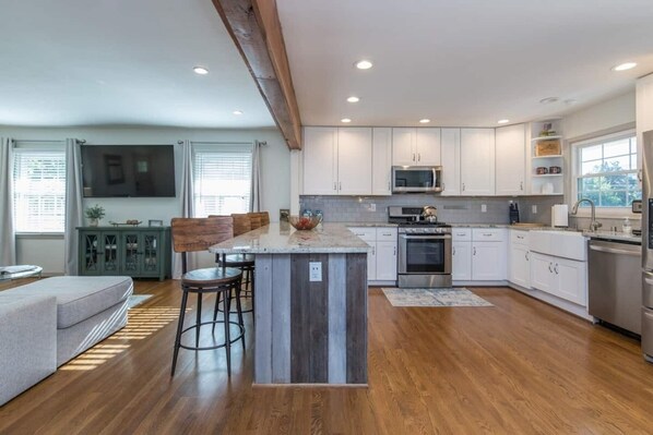 A modern kitchen with white cabinets, a central island, and a dining area with a grey sofa and wooden stools.