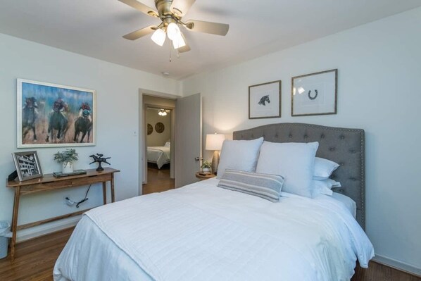 A bedroom with a large bed, a ceiling fan, a bedside table with a lamp, and a painting on the wall.