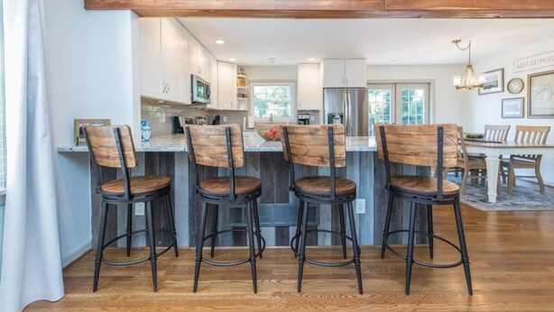 A modern kitchen with wooden bar stools, a dining area with a chandelier, and a large wooden beam ceiling.