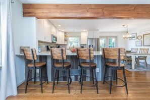 A modern kitchen with wooden bar stools, a dining area with a chandelier, and a large wooden beam ceiling.