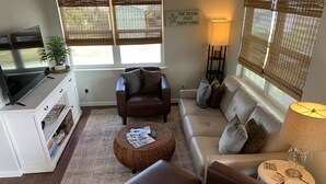 Ein Wohnzimmer mit braunem Ledersofa, einer weißen Unterhaltungseinheit, einem Korbtisch und einem großen Fenster mit Jalousien.