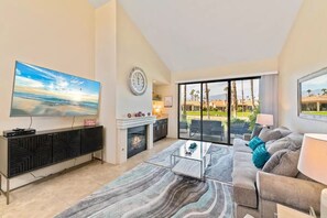 A spacious living room with a large flat-screen TV, a fireplace, a sofa, and a view of the outdoors. - Spectacular golf and water views! New furnishings! (Palm Desert)
