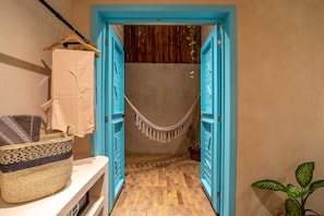 Un couloir étroit, avec des portes bleues, mène à une pièce. Un panier en osier est posé sur une étagère blanche, et une plante en pot est visible sur la droite.