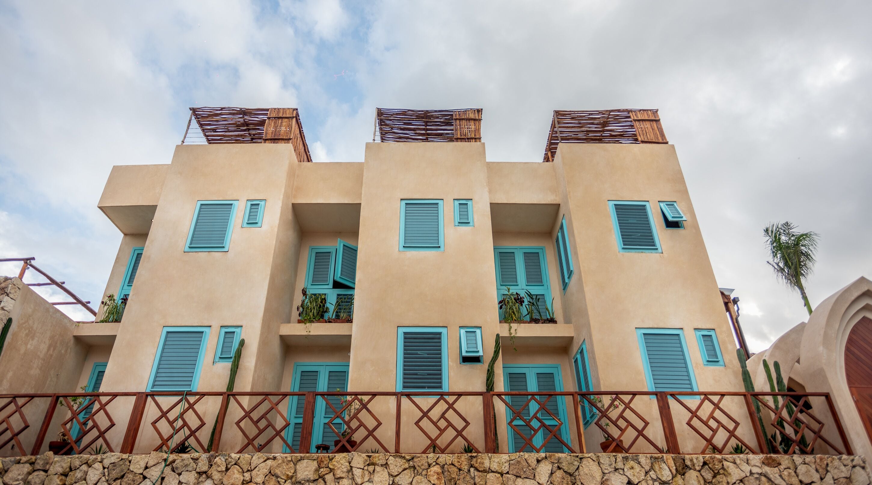 Un edificio de varios pisos con balcones, un muro de piedra al frente y un cielo despejado.