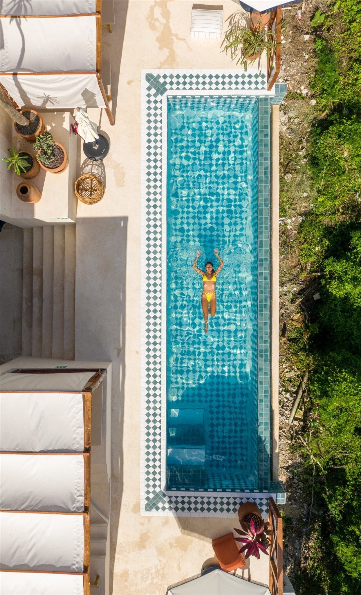 Vista aérea de una piscina con borde de azulejos, rodeada de sillones de descanso y plantas en macetas.