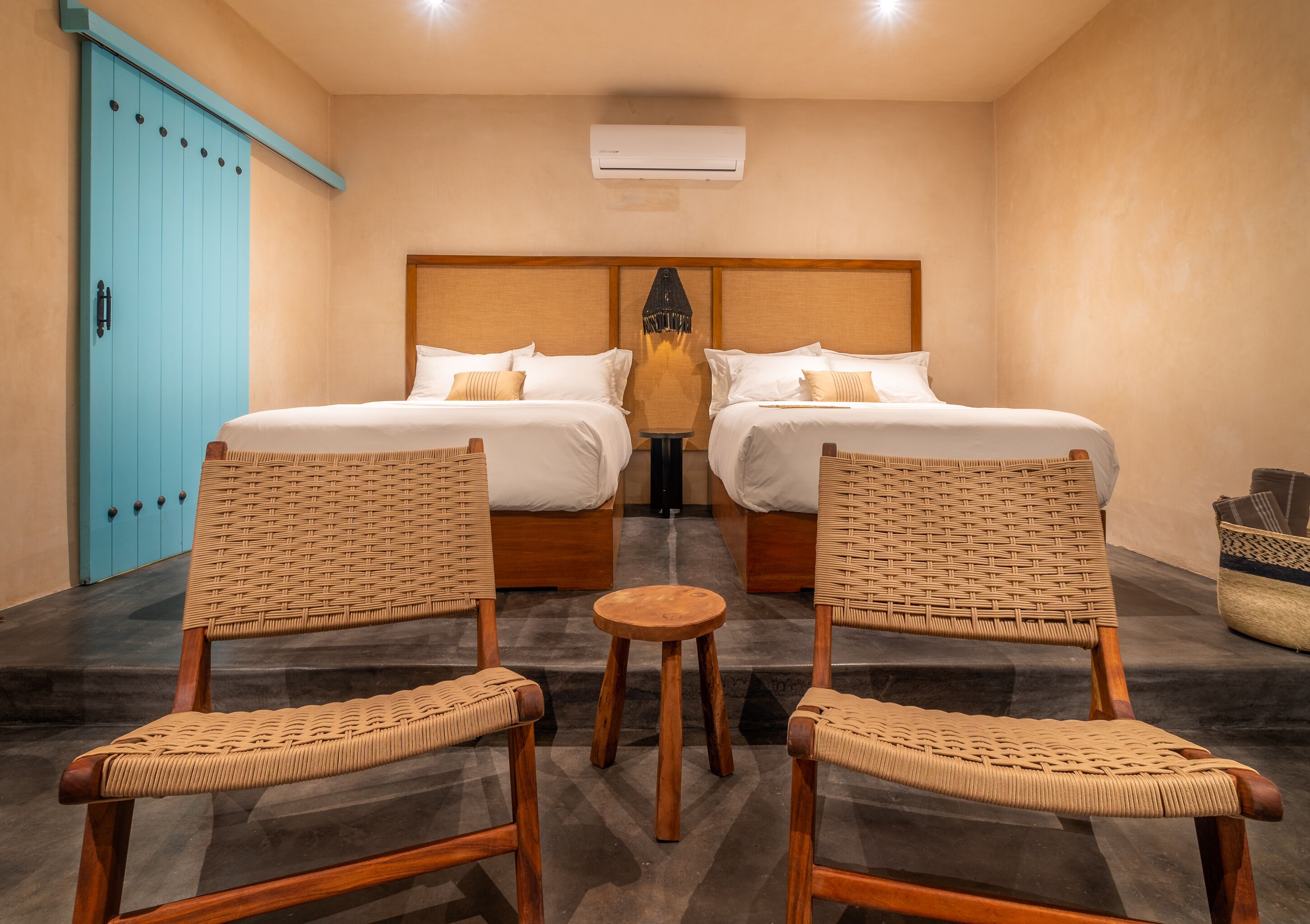 A hotel room with two beds, a small round table, two wicker chairs, a blue wardrobe, and a ceiling air conditioner.