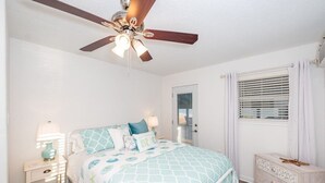 Un dormitorio con cama, mesitas de noche, cómoda, ventilador de techo y ventana con persianas.