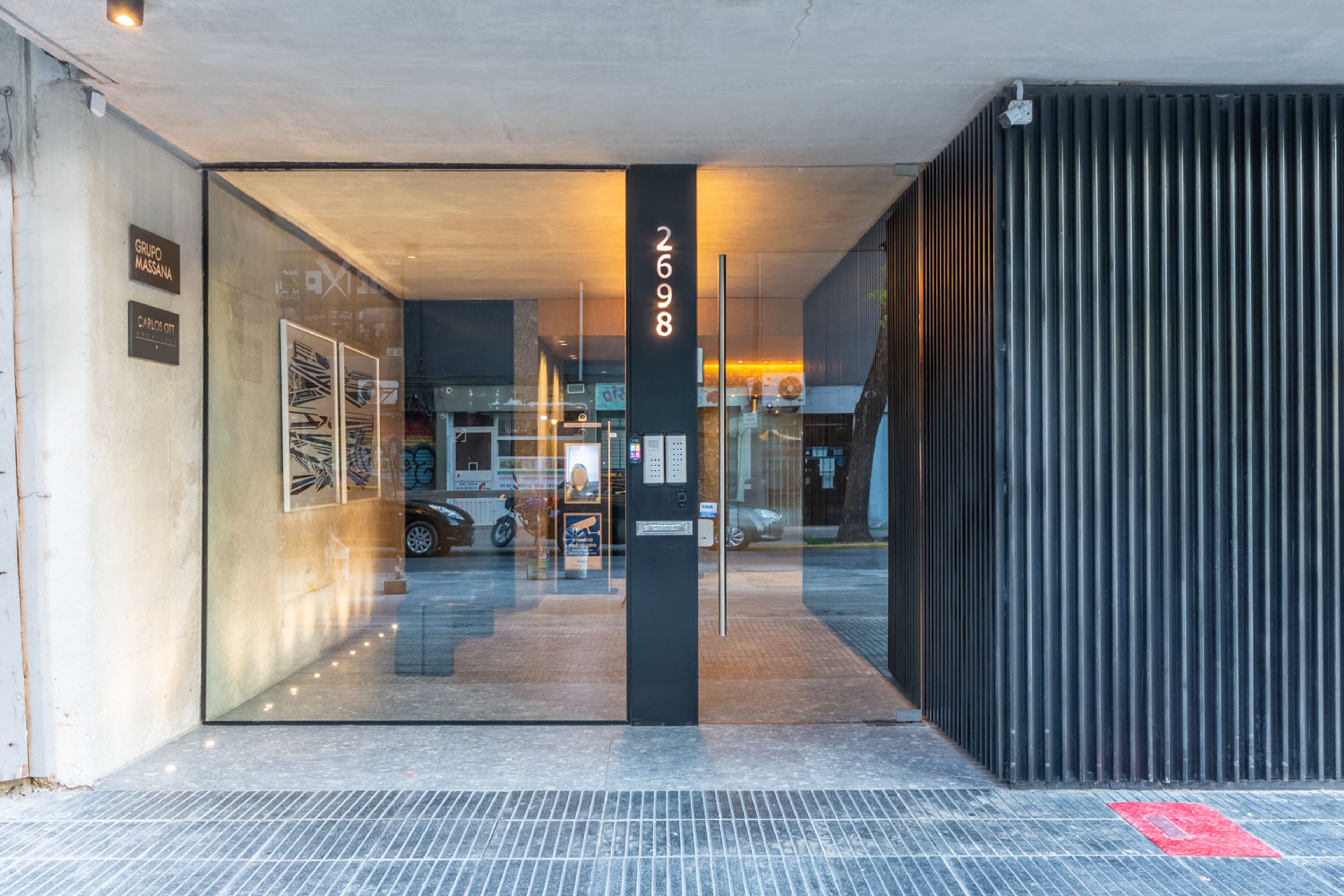 Una entrada moderna a un edificio con fachada de vidrio, un pilar negro con el número 2698 y un camino pavimentado táctil de color rojo.