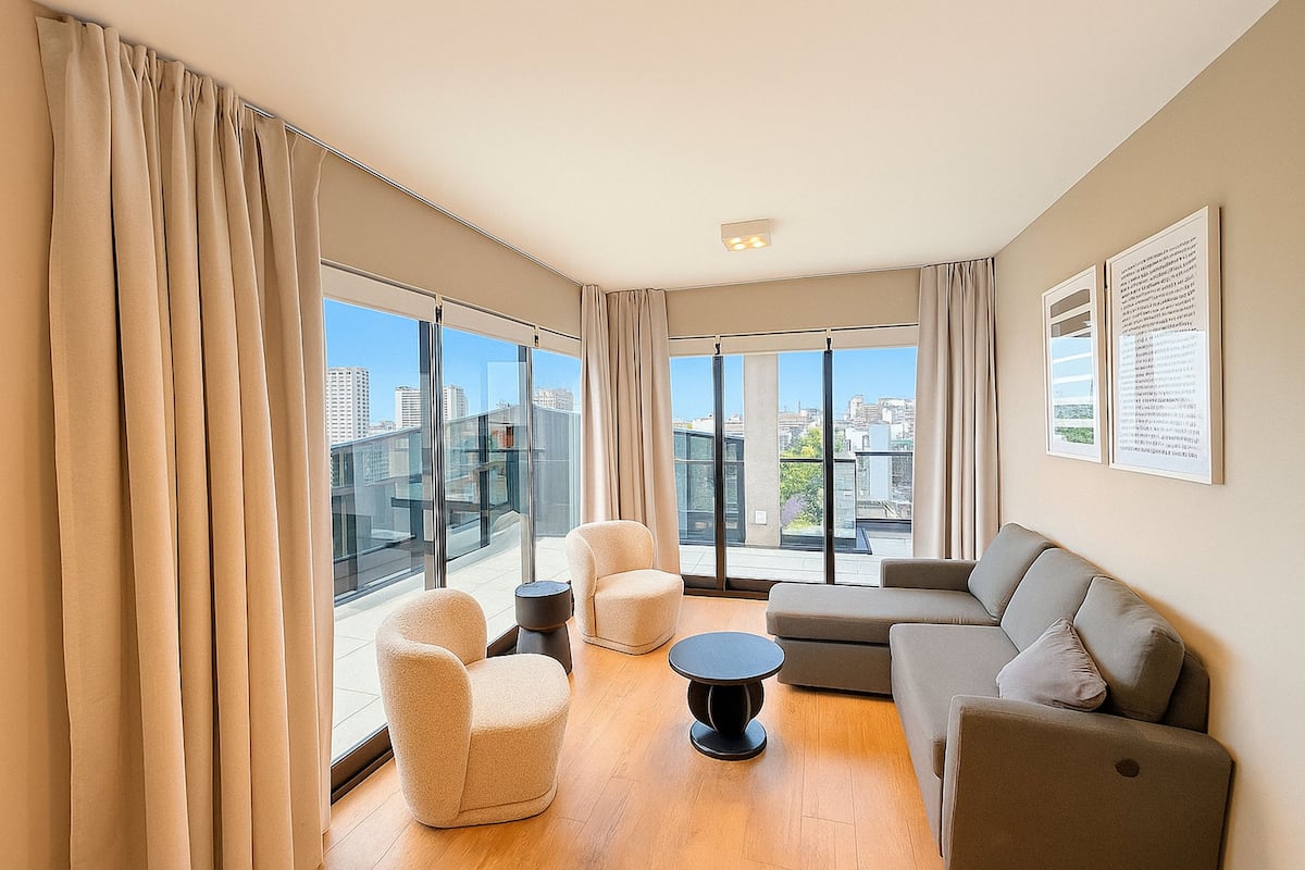 A modern living room with large windows, a sofa, a round table, and two armchairs.