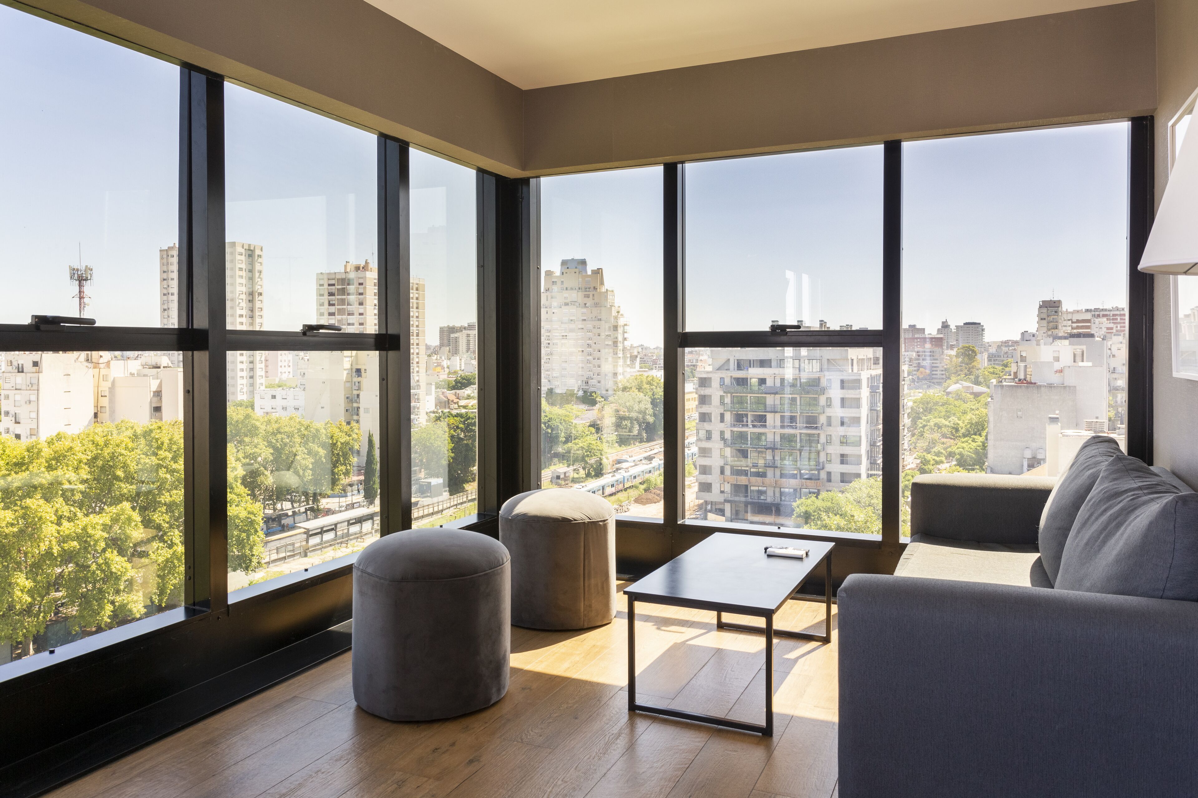 Un salon moderne avec de grandes fenêtres, un canapé gris, une petite table et deux poufs.