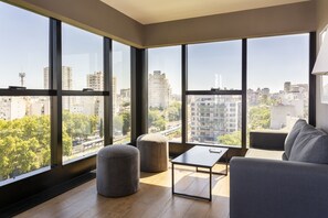 A modern living room with large windows, a grey sofa, a small table, and two ottomans. - Live Belgrano Boutique Apartments (Buenos Aires)