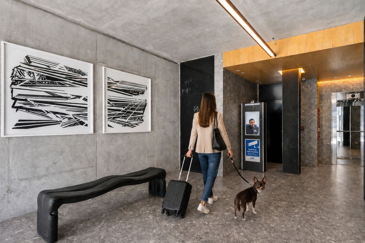 A woman with a suitcase and a dog walking in a hallway with artwork on the walls and an elevator.
