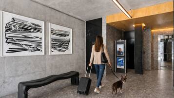 A woman with a suitcase and a dog walking in a hallway with artwork on the walls and an elevator.