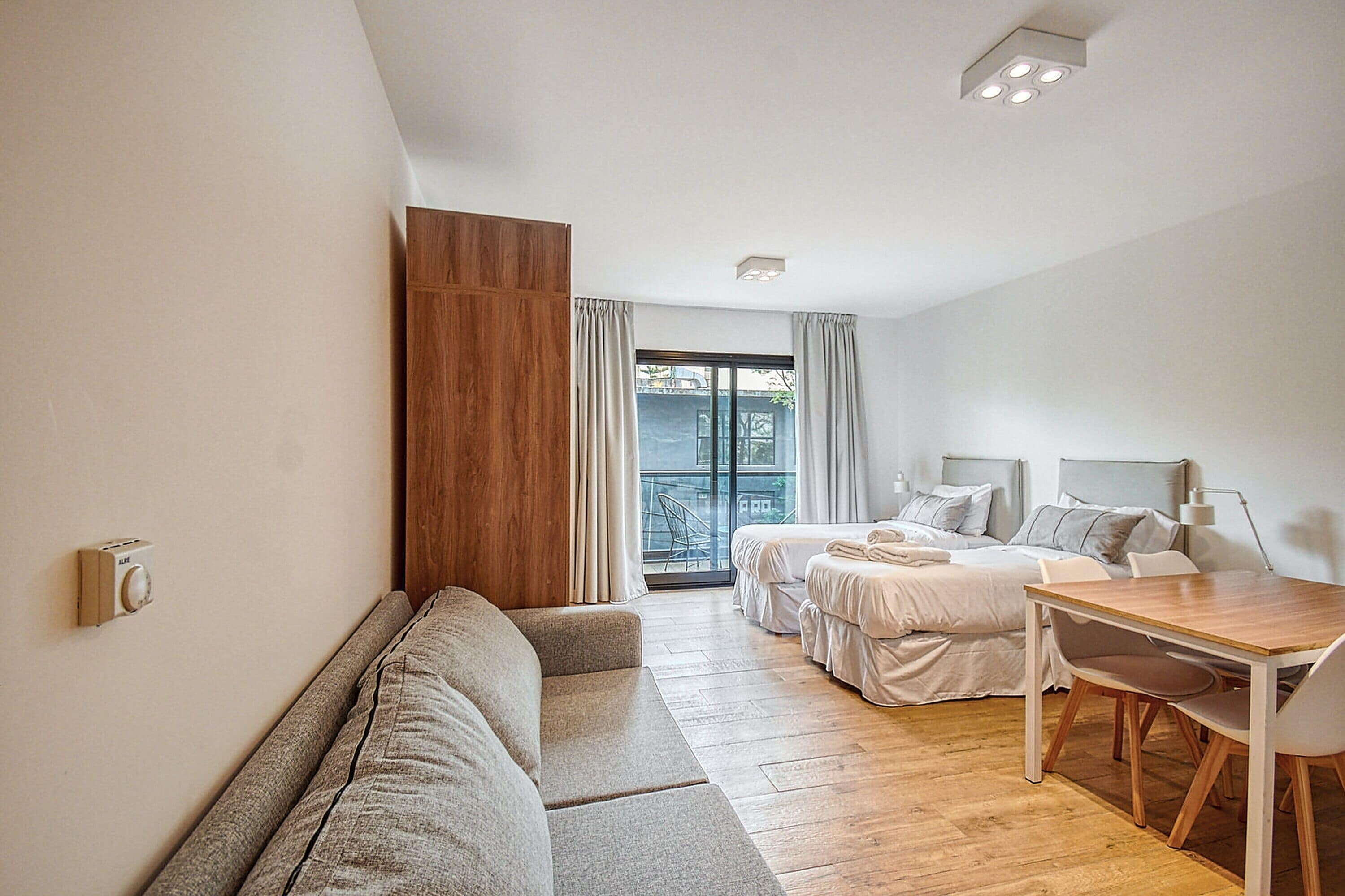A modern hotel room with a sofa, a bed, a dining table, and a balcony with a view.