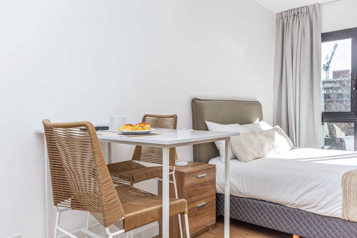 A hotel room with a bed, a desk with a chair, a small table with a plate of food, and a window with curtains.