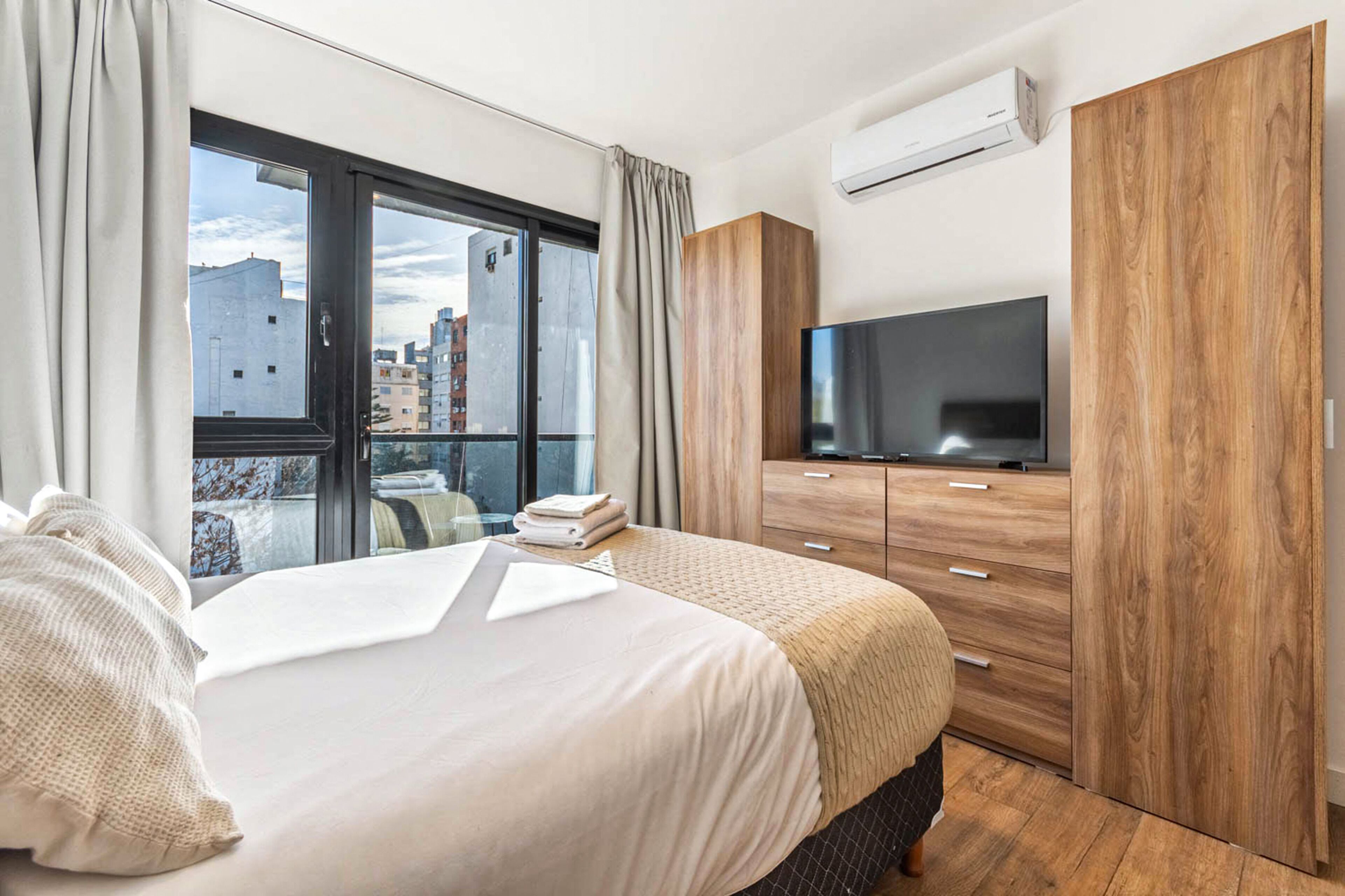 A modern hotel room with a large bed, a flat-screen TV, wooden wardrobe, and a city view.