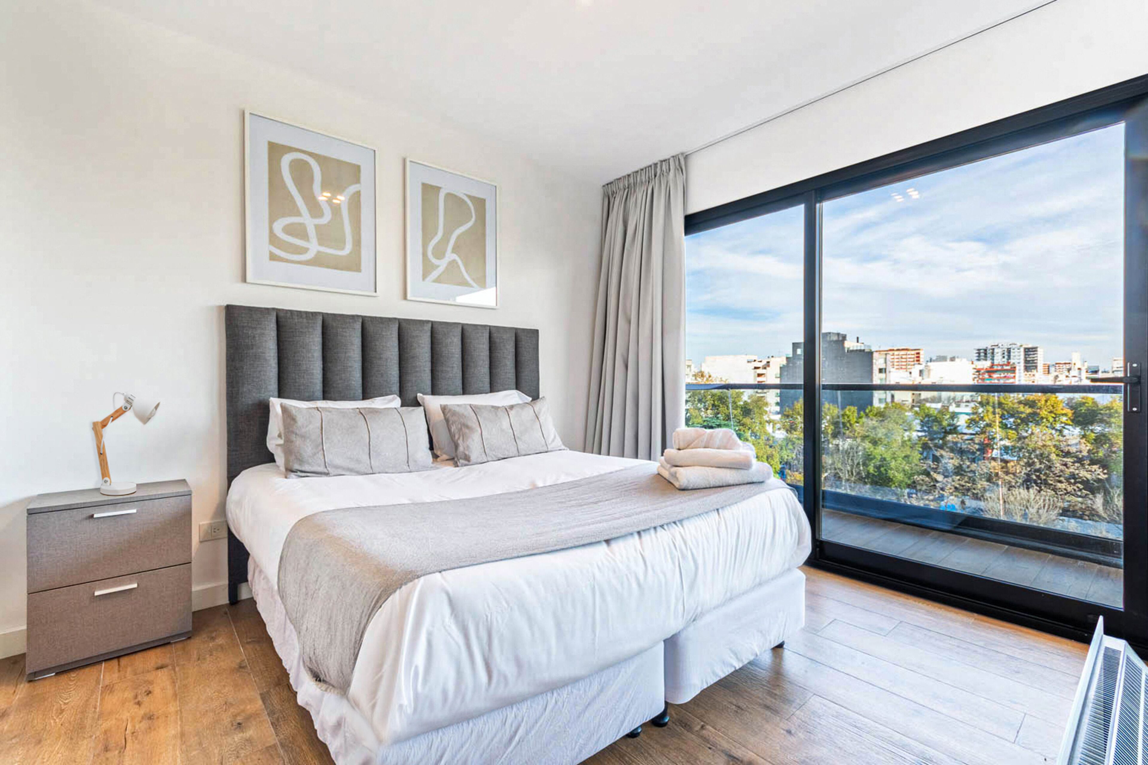 A modern bedroom with a large bed, a nightstand, and a view of the city through a large window.