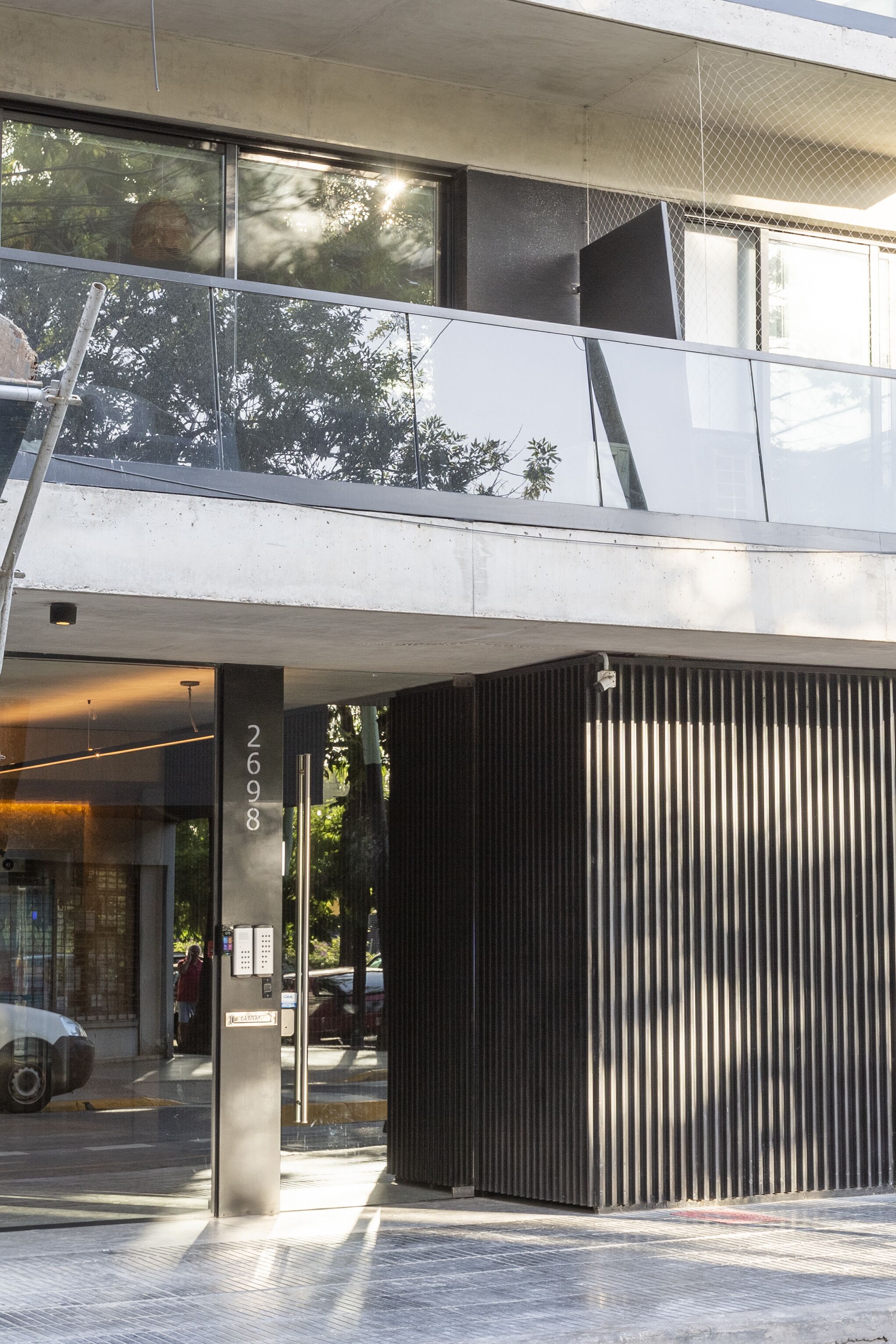 Entrada de un edificio moderno con puerta de vidrio, placa con número de metal y barandilla metálica en la parte superior.