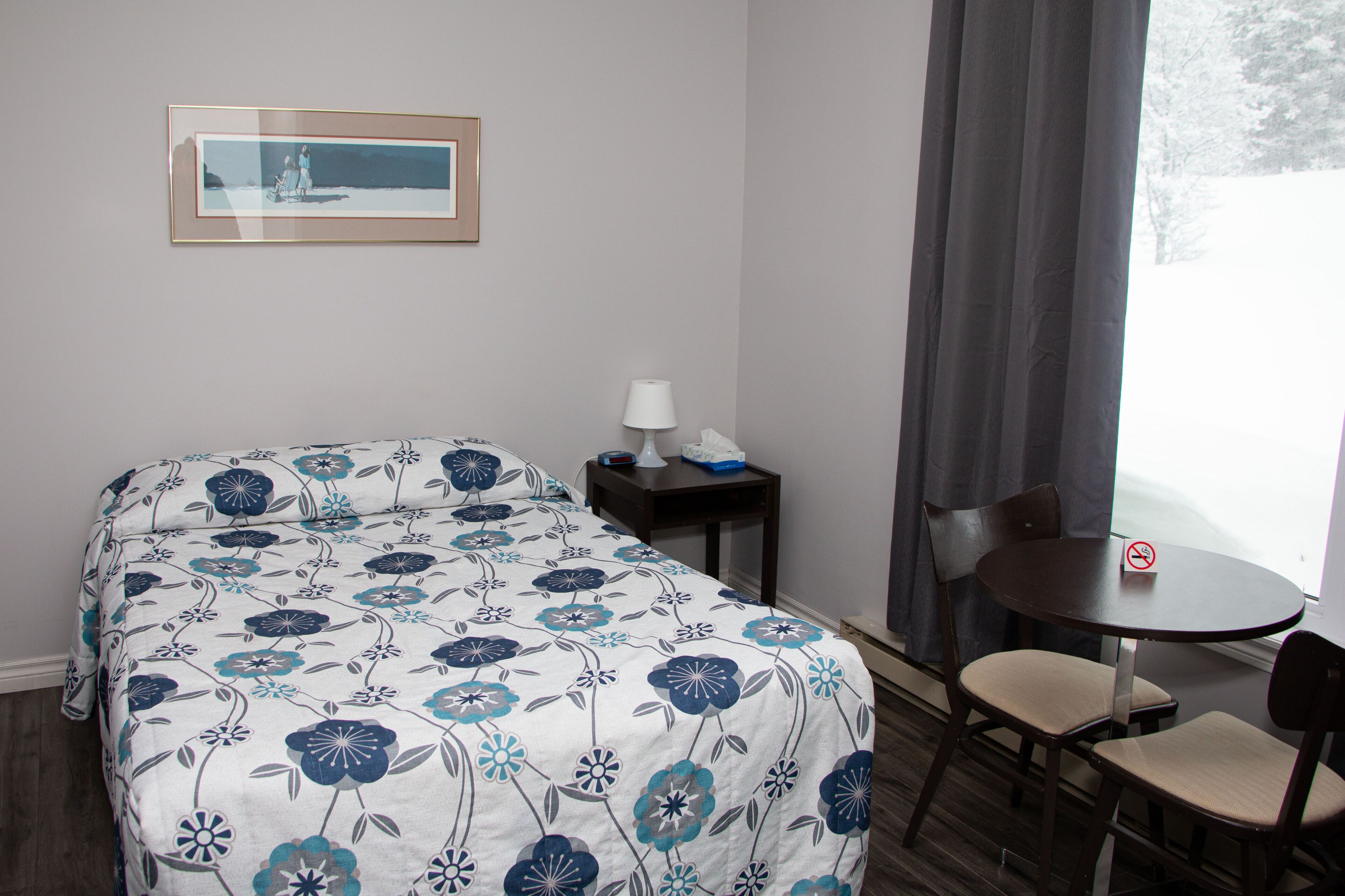 A hotel room with a bed, a small table with a lamp, a chair, and a framed picture on the wall.