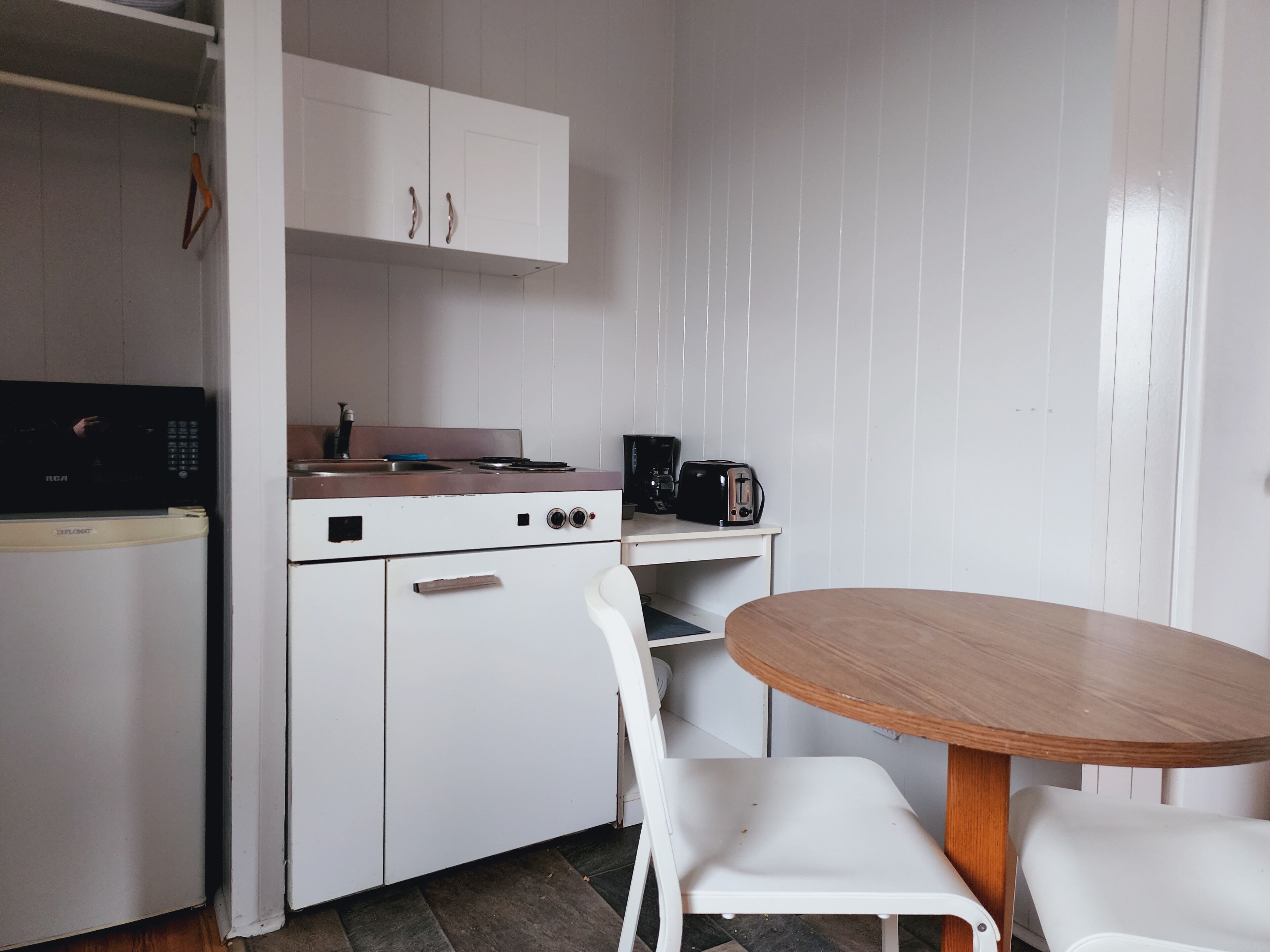 A compact kitchen with white cabinets, a stove, a microwave, and a coffee maker. There is a small table with two white chairs.