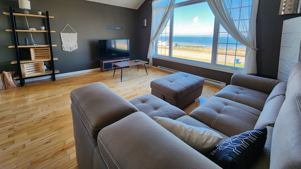 Una sala de estar moderna con un ventanal grande, un televisor de pantalla plana, piso de madera y pared oscura.