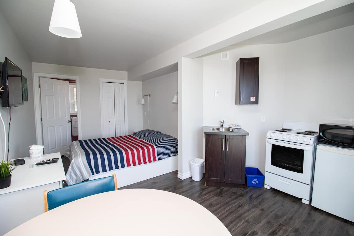A compact room with a bed, a small kitchenette, a desk with a chair, and a TV mounted on the wall.