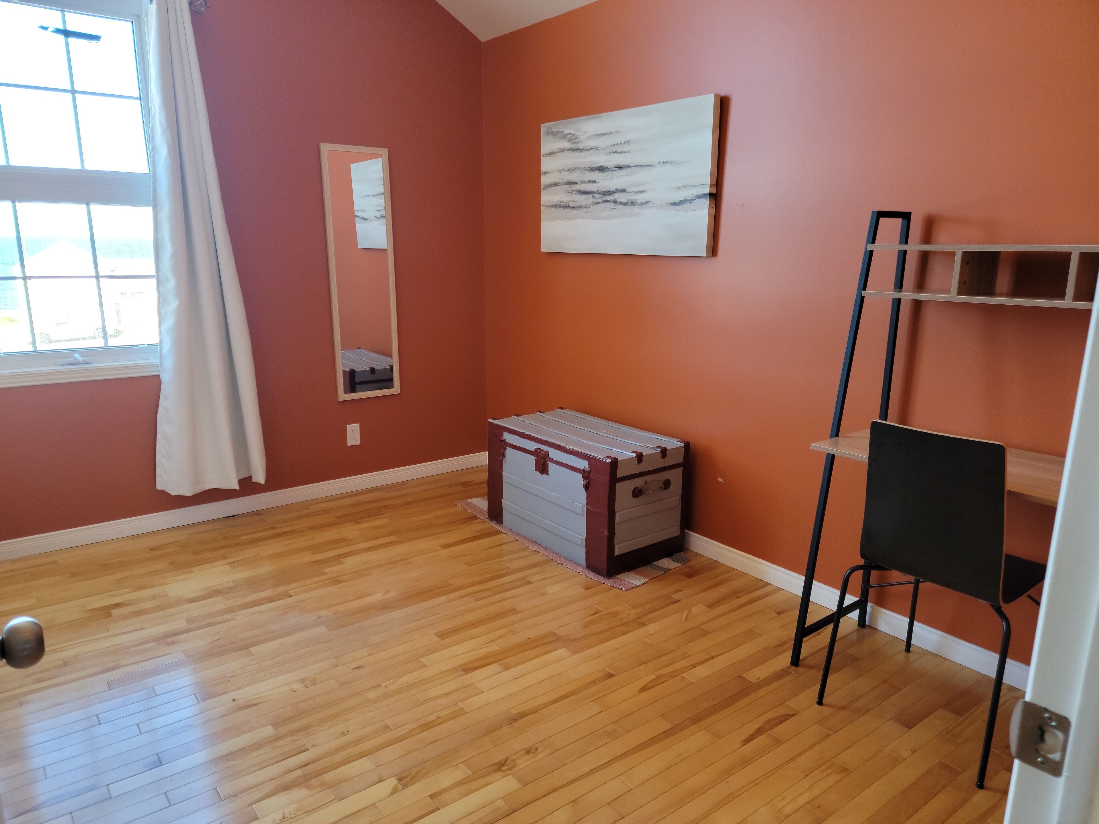Ein Zimmer mit Holzboden, einem weißen Schminktisch, einem schwarzen Stuhl, einem Bild an der Wand und einem Fenster mit weißen Vorhängen.