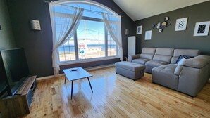 Un salón con un ventanal grande, un sofá gris, una mesita y un televisor.