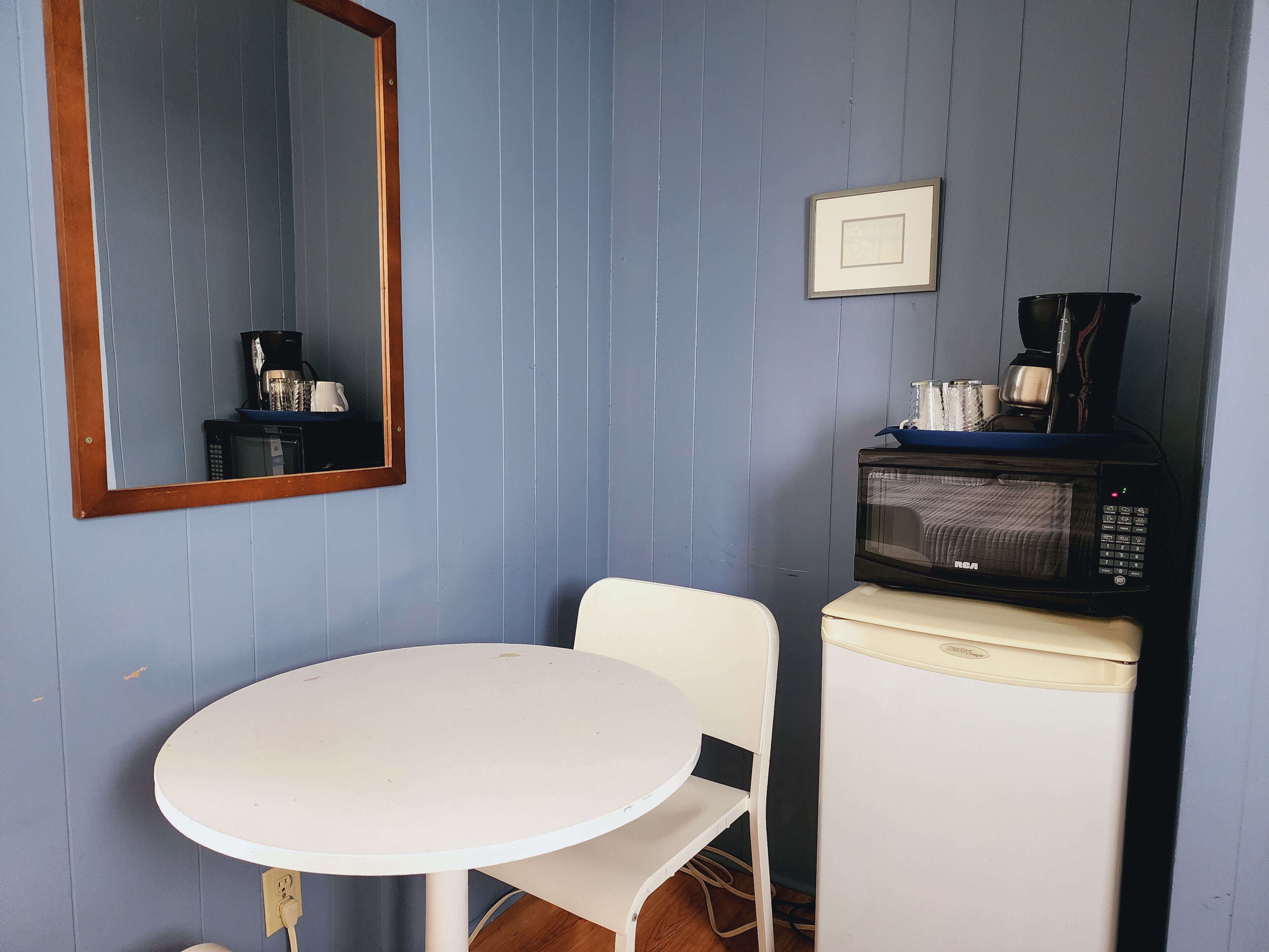 Een kleine kamer met een witte tafel en stoel, een magnetron, een koffiezetapparaat en een lijst aan de muur.