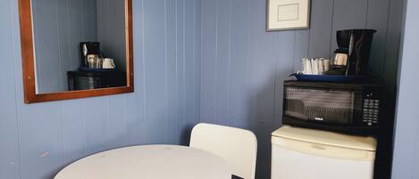 Een kleine kamer met een witte tafel en stoel, een magnetron, een koffiezetapparaat en een lijst aan de muur.