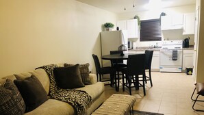 Una sala de estar con un sofá, una mesa de centro y un comedor con sillas y una mesa. Se observa una cocina al fondo con armarios y electrodomésticos.